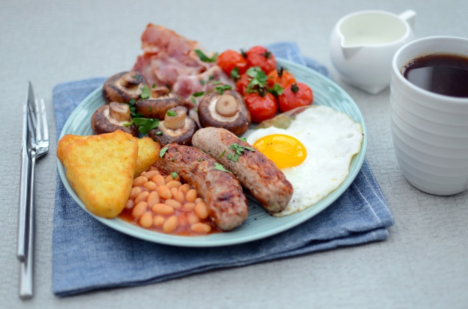A traditional Irish breakfast on a blue napkin, including crispy rashers, pork sausages, fried egg, grilled tomatoes, sautéed mushrooms and baked beans, alongside a white mug of black coffee.