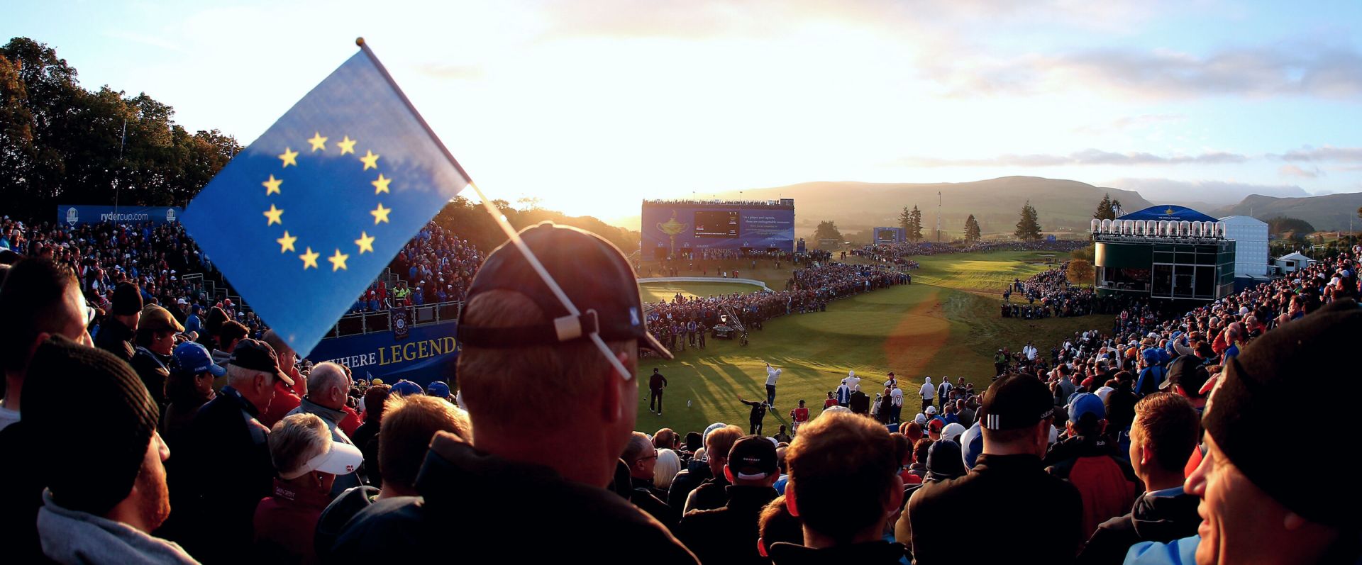 Crowd at a golf event with European flag and sunset in the background