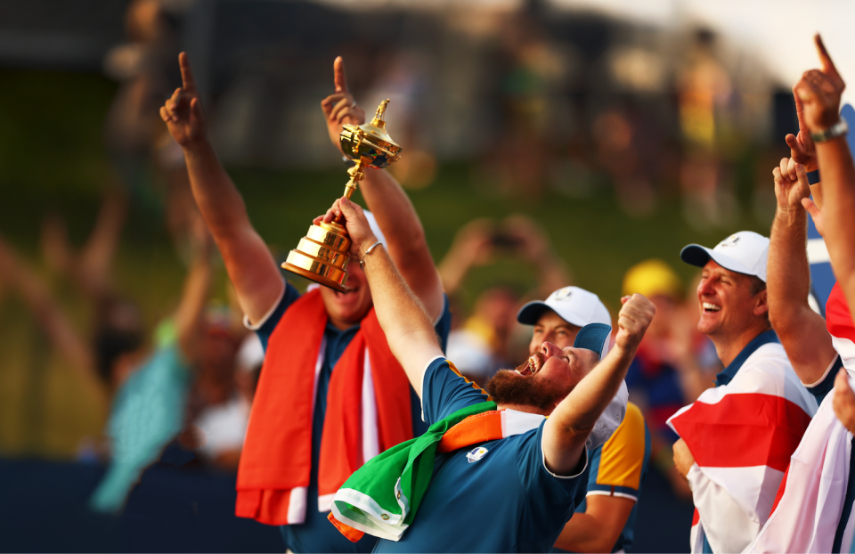 A group of men with colourful flags around their shoulders, celebrating a victory with one man holding a gold trophy.